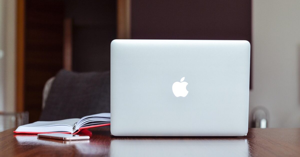 アップルのロゴが光るMacBookと開いたノートとスマートフォンが置かれたデスク
