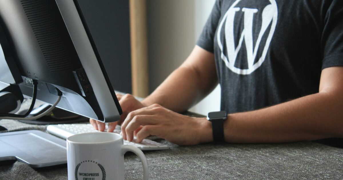 WordPressのロゴが入ったTシャツを着た人物がデスクでパソコン作業をしている様子